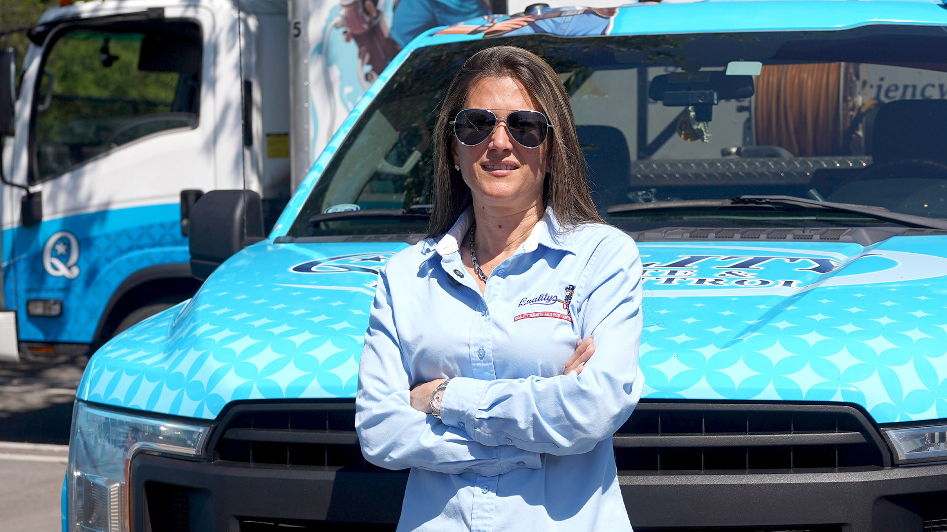 Woman in light blue uniform with folded arms standing in front of a blue service truck with graphics and logo, wearing sunglasses.